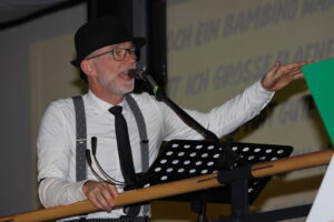 Ein Mann mit Hut und Krawatte spricht in ein Mikrofon, vor einem Notenständer stehend.