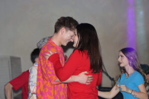 Zwei Personen tanzen eng beieinander, lächelnd. Im Hintergrund eine weitere Person in Blau.