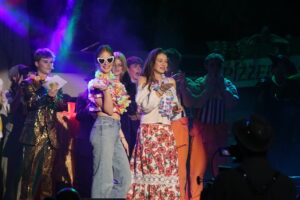 Bunt gekleidete Personen mit Herzbrillen und Blumenketten auf einer Bühne. Ein Mann im goldenen Anzug applaudiert.