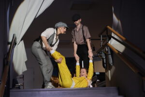 Drei Personen auf einer Bühne, zwei ziehen eine Person im gelben Outfit über eine Stufe.