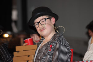 Jemand in gestreifter Jacke und Hut mit Mikrofon trägt eine rote Schleife, hält Pizzakartons und eine rote Tasse.