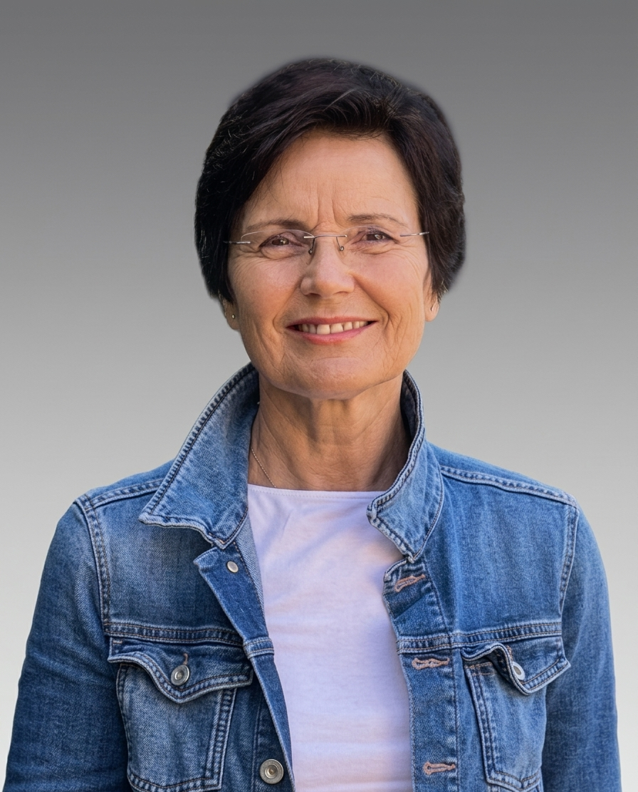 Eine lächelnde Frau mit kurzer dunkler Frisur, Brille und Jeansjacke vor grauem Hintergrund.