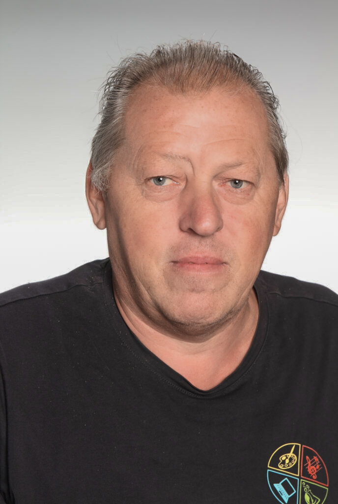 Mann mit grauem Haar, schwarzem T-Shirt und neutralem Gesichtsausdruck vor grauem Hintergrund.