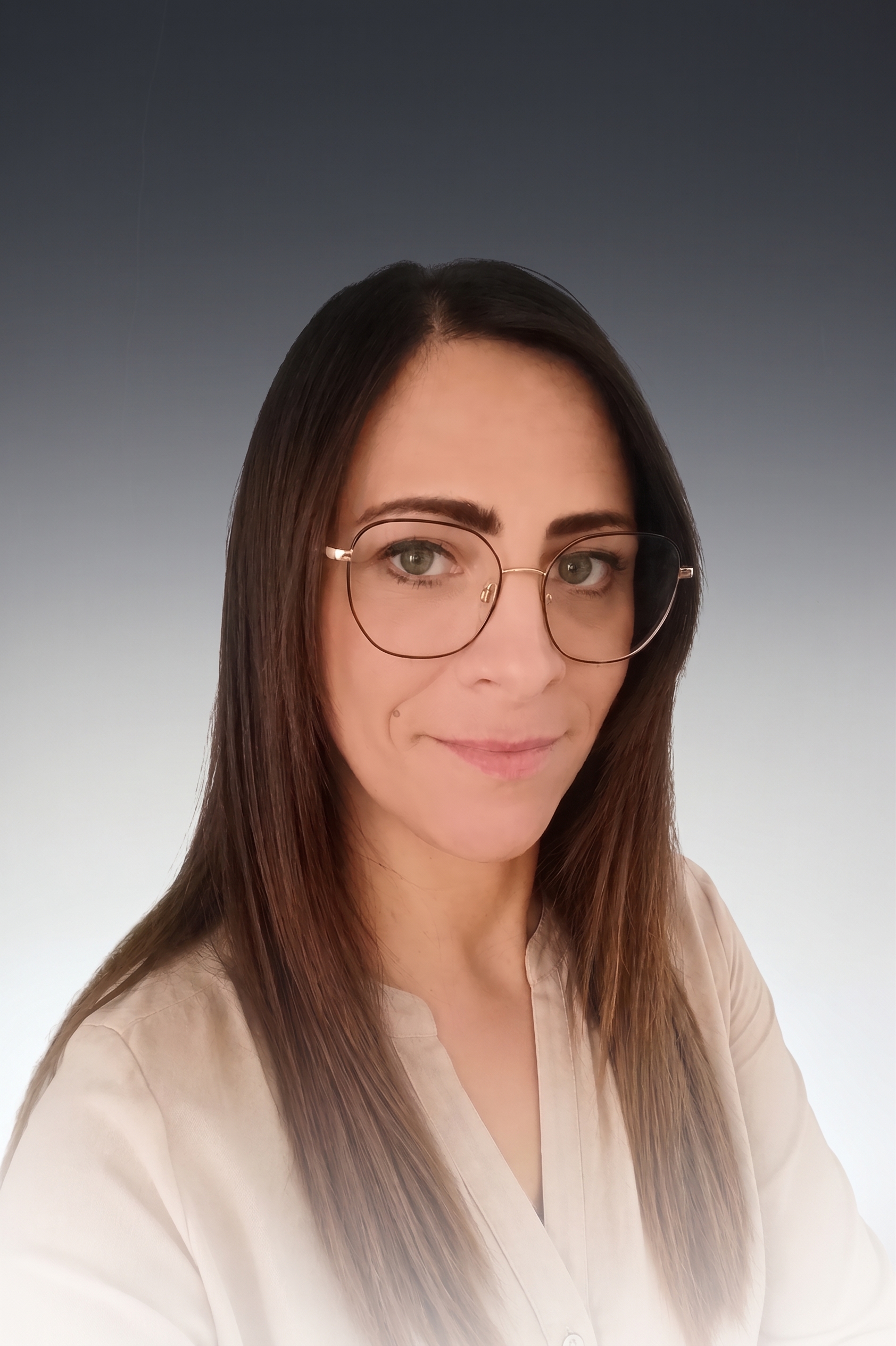 Frau mit Brille und langen braunen Haaren, trägt beigefarbenes Oberteil, lächelt leicht; grauer Hintergrund.